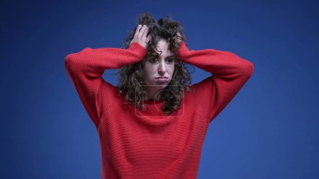 Téléchargez les photos : Frustré jeune femme tirant ses cheveux dans l'anxiété et le désespoir sur fond bleu avec pull rouge. Anxieux 20s personne féminine - en image libre de droit