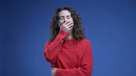 Tired woman yawning covering mouth with hand feeling fatigue and boredom standing on blue backdrop