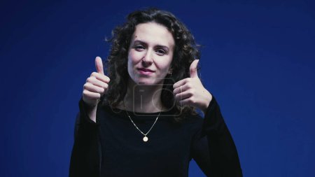 Photo for Positive Young Woman Giving Thumbs Up, Blue Background Signifying Approval - Royalty Free Image