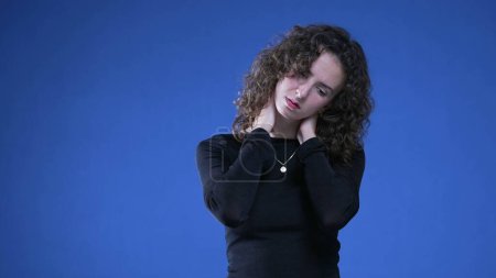 Foto de Mujer joven estresada frotando el cuello tratando de apaciguar la angustia mental mientras está de pie sobre fondo azul - Imagen libre de derechos
