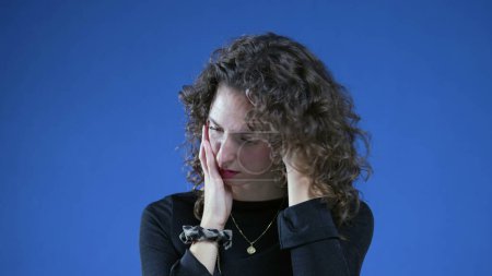 Téléchargez les photos : Femme anxieuse dans la vingtaine se sentant nerveuse et remplie d'anxiété debout en arrière-plan bleu avec regard introspectif coûteux - en image libre de droit