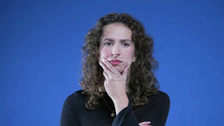Photo for Pensive woman thinking deeply with hand on chin standing on blue background. Thoughtful expression of a caucasian female person in 20s with curly hair - Royalty Free Image