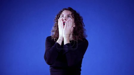 Foto de Mujer ansiosa reaccionando a las noticias con conmoción y desesperación sobre fondo azul, cubriendo la boca con las manos - Imagen libre de derechos