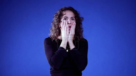 Foto de Mujer ansiosa reaccionando a las noticias con conmoción y desesperación sobre fondo azul, cubriendo la boca con las manos - Imagen libre de derechos