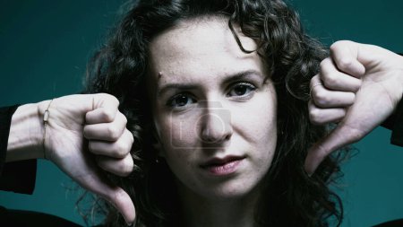 Photo for Woman Giving Thumbs Down in Rejection, Close-up of Female in 20s with Negative Gesture behind teal background - Royalty Free Image