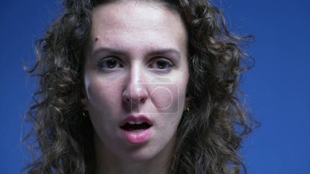 Photo for Scared woman reacts with shock and unbelief by covering mouth feeling stress and preocupation, looking directly at camera with blue background. Close-up face 20s person - Royalty Free Image