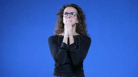 Photo for Preoccupied young woman standing on blue background looking up with hands clenched together holding her breath during stressful times - Royalty Free Image
