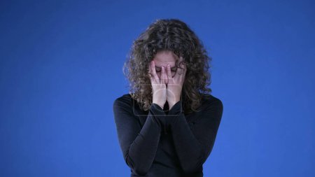 Téléchargez les photos : Femme frustrée éprouvant des regrets et de la confusion au sujet des souvenirs passés, femme dans la vingtaine couvrant le visage de honte et d'anxiété debout sur fond bleu - en image libre de droit