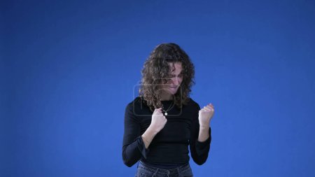 Photo for Happy woman celebrating success by raising fists in the air looking at camera while standing on blue background - Royalty Free Image