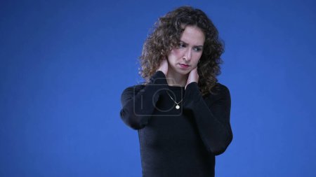 Téléchargez les photos : Stressé jeune femme frottant le cou en essayant d'apaiser l'angoisse mentale tout en se tenant debout sur fond bleu - en image libre de droit