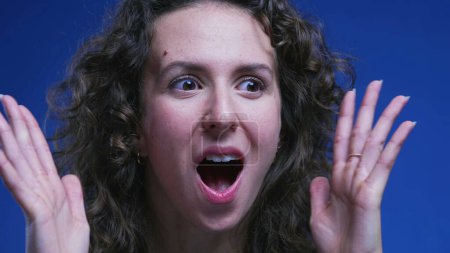 Téléchargez les photos : Femme réagissant avec choc et surprise, le visage en gros plan d'une femme dans la vingtaine stupéfaite se sentant étonnée de nouvelles sur fond bleu - en image libre de droit