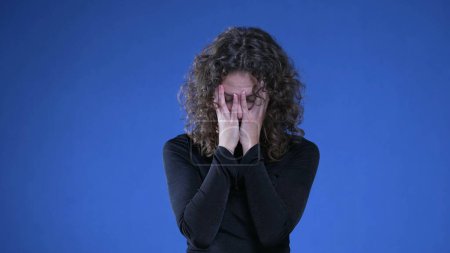 Téléchargez les photos : Femme frustrée éprouvant des regrets et de la confusion au sujet des souvenirs passés, femme dans la vingtaine couvrant le visage de honte et d'anxiété debout sur fond bleu - en image libre de droit