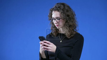 Téléchargez les photos : Femme utilisant un appareil de téléphone portable tout en se tenant debout sur fond bleu. Femme dans la vingtaine portant des lunettes de lecture texto message au téléphone avec expression neutre - en image libre de droit