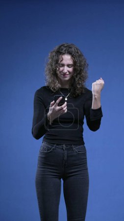 Téléchargez les photos : Femme tirant appareil de téléphone portable et célèbre de grandes nouvelles sur la notification d'écran de téléphone, debout sur fond bleu - en image libre de droit