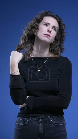 Foto de Mujer pensativa juega con el pelo mientras reflexiona decisión, soñando despierto foto de 20s persona en profunda introspección de pie sobre fondo azul - Imagen libre de derechos