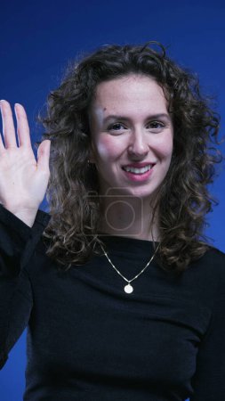 Photo for Joyful woman waves HELLO to camera on blue backdrop. One person in 20s saying HI - Royalty Free Image