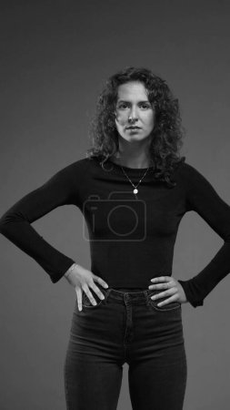 Photo for Stern serious woman looking at camera feeling angry, puts hands on hips looking directly at camera in black and white - Royalty Free Image