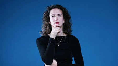 Photo for Woman contemplative decision making while squinting and with hand in chin pondering solution gazing at distance on blue background - Royalty Free Image