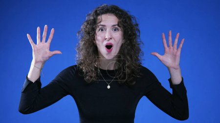Photo for Surprised woman reacting with disbelief to camera raising arms and saying "WOW" captured in slow-motion standing on blue background - Royalty Free Image
