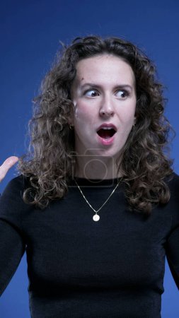 Photo for Young woman surprised reaction in SHOCK and DISBELIEF looking at camera on blue backdrop - Royalty Free Image