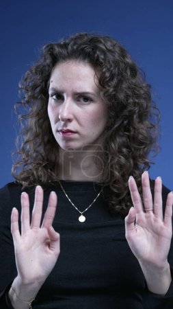 Photo for Unhappy woman rejecting offer by saying NO with body language, making an X with arms and waving finger in negation, on blue backdrop - Royalty Free Image