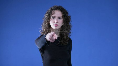 Téléchargez les photos : Femme accusant spectateur en pointant du doigt la caméra tout en se tenant sur fond bleu. Agacé personne douteuse gestuelle au délinquant - en image libre de droit