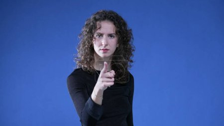 Photo for Woman accusing viewer by pointing finger at camera while standing on blue background. Upset doubtful person gesturing at offender - Royalty Free Image