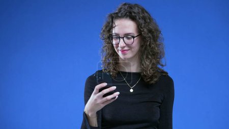 Téléchargez les photos : Femme aimant la notification de message sur le téléphone portable. femme dans la vingtaine portant des lunettes de lecture tenant le téléphone et souriant aux nouvelles positives - en image libre de droit