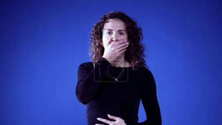 Foto de Mujer reaccionando a noticias impactantes con la mano sobre la boca sobre fondo azul, incredulidad en sus ojos - Imagen libre de derechos