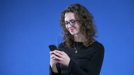 Téléchargez les photos : Femme recevant un message positif inattendu dispositif de notification de téléphone portable tout en se tenant debout sur fond bleu sourire - en image libre de droit