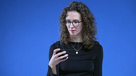 Photo for Woman frowning to message notification while holding cellphone device on blue background. Person wearing glasses reacting to unexpected news - Royalty Free Image