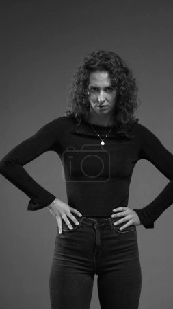Photo for Stern serious woman looking at camera feeling angry, puts hands on hips looking directly at camera in black and white - Royalty Free Image