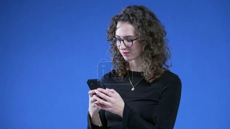 Foto de Mujer recibiendo inesperada notificación de mensaje positivo dispositivo de teléfono móvil mientras está de pie sobre fondo azul sonriendo - Imagen libre de derechos