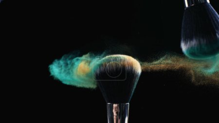 Foto de Cepillo de maquillaje que toca con partículas cosméticas en cámara súper lenta sobre fondo oscuro - Imagen libre de derechos