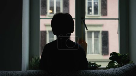 Photo for Silhouette of child staring at snow fall from home window during winter season. Back of young boy hypnotized by nature during moody day, December season - Royalty Free Image