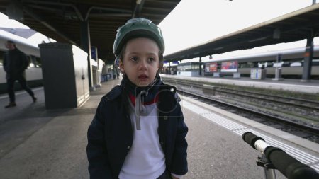 Téléchargez les photos : Pensive Boy avec casque rêver à la plate-forme de train - En attendant que le transport arrive, porter une veste et une écharpe pendant la saison d'automne - en image libre de droit