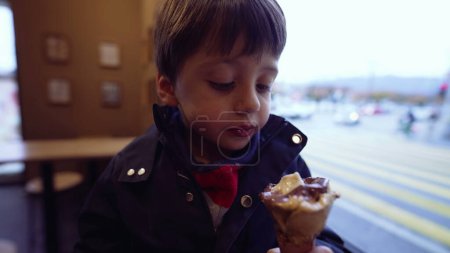 Téléchargez les photos : Jeune enfant mangeant de la crème glacée avec paysage urbain en arrière-plan, garçon envoûtant friandise dessert sucré au salon de crème glacée - en image libre de droit