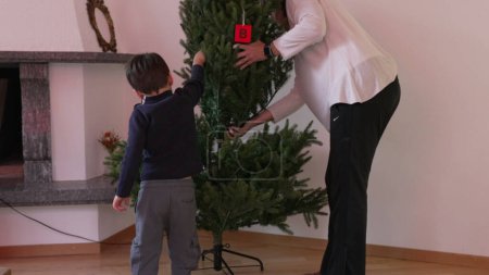 Téléchargez les photos : Mère et enfant installant le sapin de Noël pendant la saison des fêtes dans le salon par cheminée - en image libre de droit