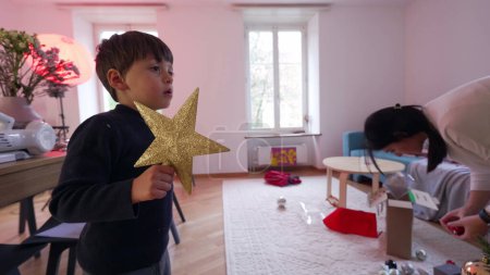 Photo for Child holding star eagerly waiting to put it on top of Christmas tree during winter holiday season, family preparing to festivities decorating tree by adding ornamentation - Royalty Free Image