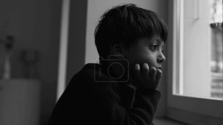 Foto de Niño deprimido se para junto a la ventana mirando a la vista con expresión triste introspectiva en monocromo malhumorado, dramático blanco y negro. niño sintiendo soledad - Imagen libre de derechos