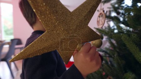 Foto de Niño sosteniendo la estrella con impaciencia esperando para ponerlo en la parte superior del árbol de Navidad durante la temporada de vacaciones de invierno, la familia se prepara para las festividades decorando el árbol mediante la adición de ornamentación - Imagen libre de derechos