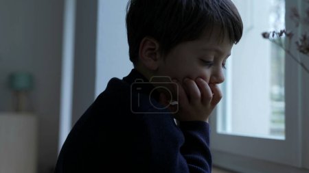 Téléchargez les photos : Petit garçon réfléchi avec la main dans le menton rêvant à la maison par la fenêtre. Pensée pénitentiaire profondément absorbée dans les pensées introspectives - en image libre de droit