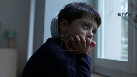 Téléchargez les photos : Petit garçon réfléchi avec la main dans le menton rêvant à la maison par la fenêtre. Pensée pénitentiaire profondément absorbée dans les pensées introspectives - en image libre de droit