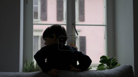 Foto de Chico joven estirando el cuerpo por la ventana mientras cae nieve afuera. Estiramiento matutino del niño durante la temporada de invierno - Imagen libre de derechos