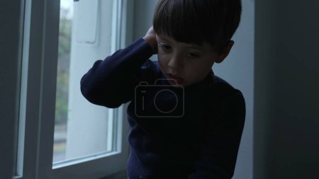 Foto de Niño aburrido atrapado en casa apoyado en la ventana del apartamento mirando a la vista desde el interior luchando con el aburrimiento - Imagen libre de derechos