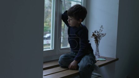 Foto de Niño sentado junto a la ventana del apartamento mirando a la vista desde casa con expresión pensativa reflexiva, niño atrapado en el interior mirando hacia fuera - Imagen libre de derechos