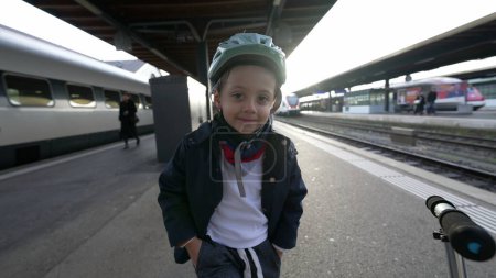Téléchargez les photos : Portrait d'un enfant heureux debout au quai de la gare souriant à la caméra tout en portant un casque à côté du scooter. Jeune garçon portant une veste souriant à la caméra - en image libre de droit