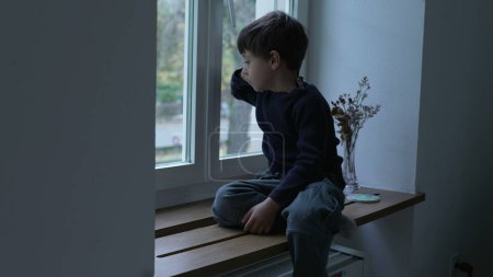 Photo for Child seated by apartment window looking out at view from home with pensive thoughtful expression, kid stuck indoors gazing out - Royalty Free Image