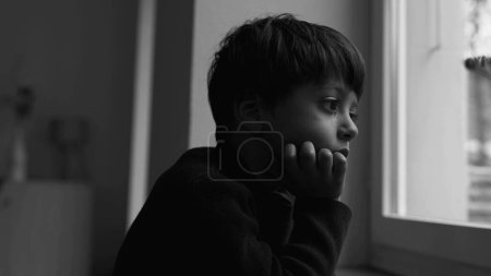 Foto de Niño deprimido se para junto a la ventana mirando a la vista con expresión triste introspectiva en monocromo malhumorado, dramático blanco y negro. niño sintiendo soledad - Imagen libre de derechos
