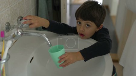 Foto de Niño bebiendo del grifo de agua en el baño usando una taza de plástico después de cepillarse los dientes durante el ritual matutino, capturando el auténtico estilo de vida familiar diario - Imagen libre de derechos